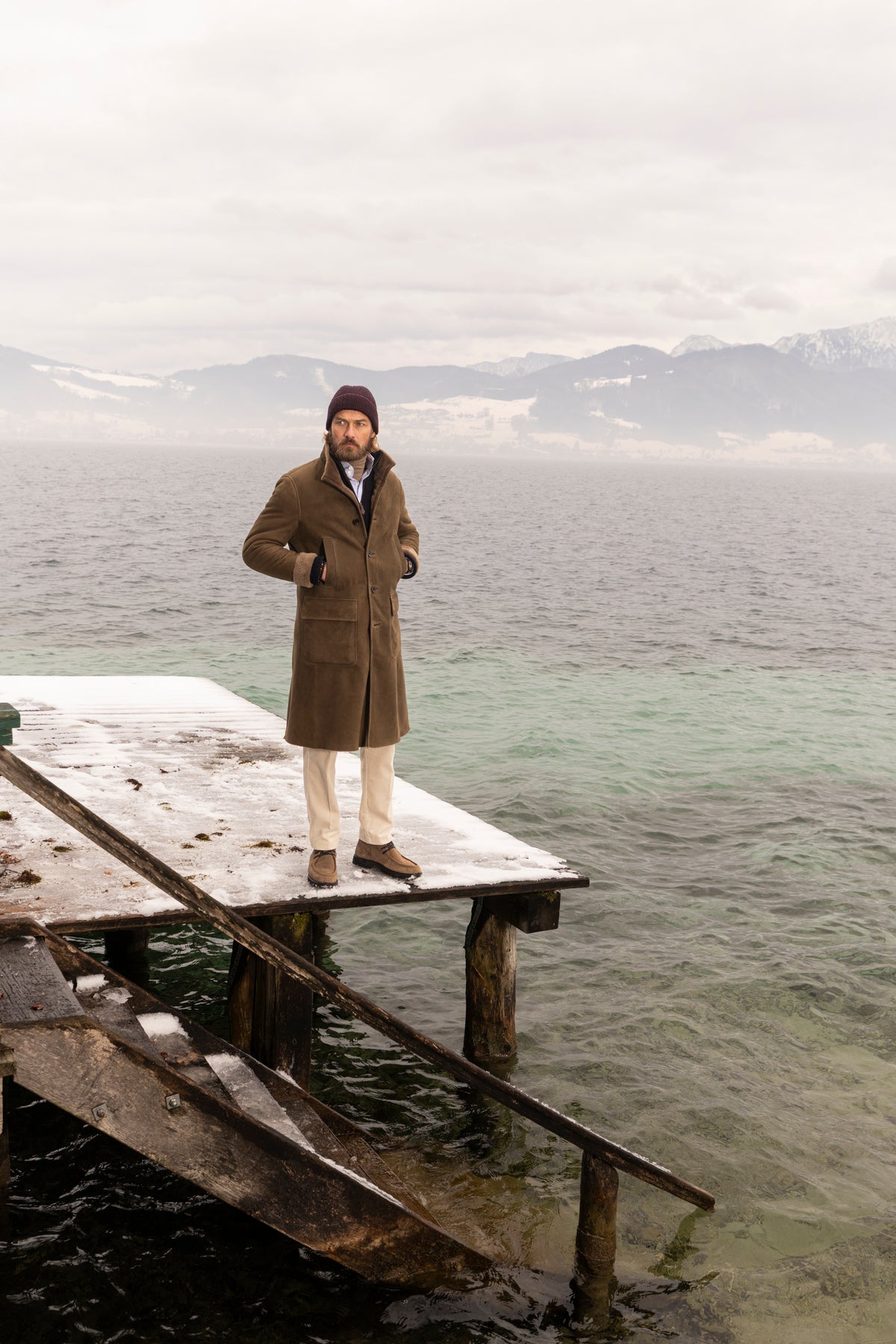 Model in Lammfellmantel auf einem verschneiten Steg, im Hintergrund winterliche Landschaft am Seeufer